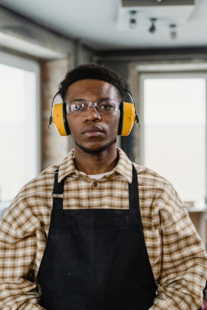 Young black craftsman wearing earmuffs and goggles in an industrial workshop.